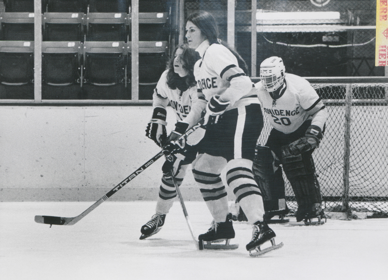 Three hockey players wearing Providence College uniforms. One stands in the goal wearing a helmet and the other two are standing on the ice with no helmets holding hockey sticks.