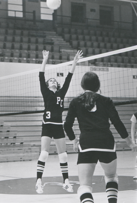 Two women wearing PC sports uniforms playing volleyball on a court in a gym. One is jumping up to hit the ball over the net.