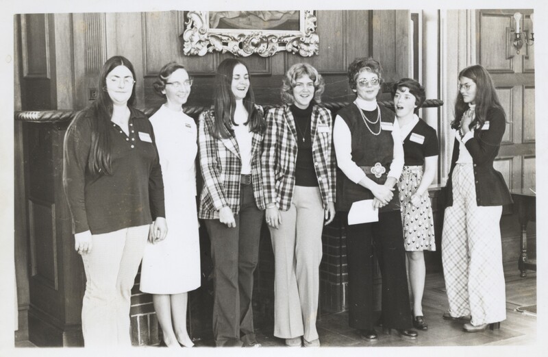Seven women standing side by side in an indoor setting with wood-paneled walls and a framed painting above them.