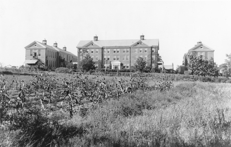 Black and white photo of a large three-story brick building with cupolas, flanked by two smaller buildings, set behind a field with rows of corn and surrounded by trees.