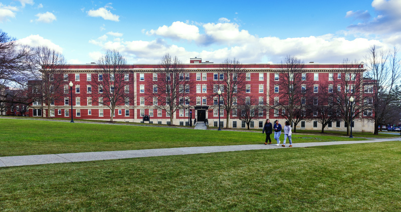 Large red-brick multi-story building with white-trimmed windows, leafless trees, and three people walking on a concrete path across a grassy lawn under a partly cloudy sky.