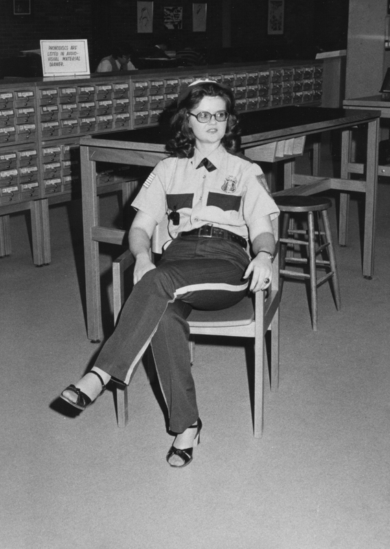 A woman wearing a security guards uniform sitting on a chair with legs crossed in the Phillips Memorial Library.