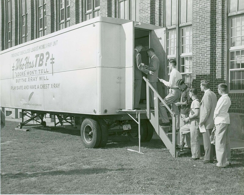 Black and white photo of a mobile X-ray unit outside a brick building, with people lined up to enter and a sign that reads 'Providence tuberculosis league's mobile x-ray unit: Who has TB? Looks won't tell but the x-ray will, play safe and have a chest x-ray.'