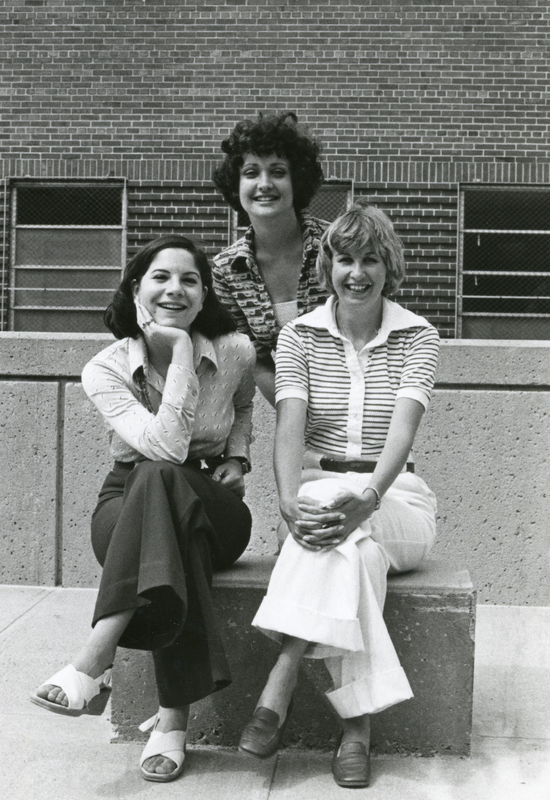Three women posing for a photograph outside. Two are sitting on a bench while the third stands behind them.