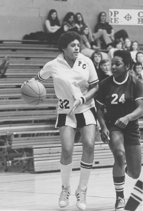 Two basketball players on the court. One has possession of the ball and is wearing a Providence College uniform. There are onlookers sitting on the bleachers in the background.
