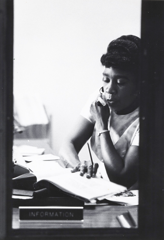 O'daylise Collins sits at a desk talking on a telephone while flipping through a notebook.