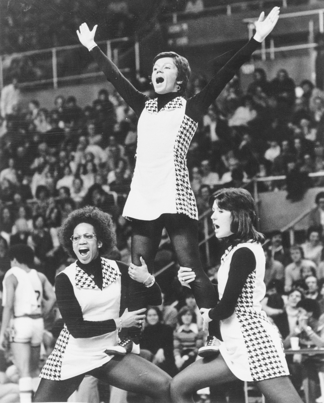 Three cheerleaders performing a stunt in front of a large crowd wearing matching uniforms with checkered patterns on the sides. Two cheerleaders are standing on the ground, supporting a third cheerleader who is elevated above them with arms raised in a 'V' shape.