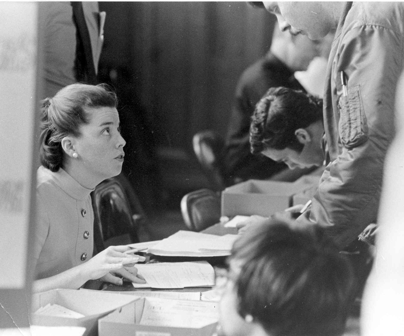 Sally J Thibodeau sitting at a registration desk with papers in front of her, interacting with a man standing on the other side of the desk.
