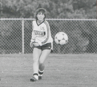 A soccer player in a Providence College uniform runs on a field with a soccer ball in the air near them.