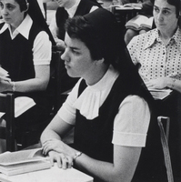 A nun sits at a desk in a classroom with a book in front of her. There are two other nuns sitting behind her.