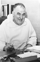 Rev. Charles V. Fennell sitting at a desk holding eyeglasses with a bookshelf in the background.