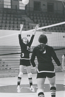 Two women wearing PC sports uniforms playing volleyball on a court in a gym. One is jumping up to hit the ball over the net.