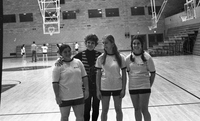 Three students in gym clothing are standing next to Helen Bert on an indoor basketball court. There are some people playing basketball in the background.