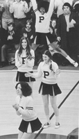 A group of cheerleaders performing at a basketball game wearing Providence College uniforms. One cheerleader is in mid-air, jumping with arms raised, while others are clapping and cheering on the court. The background shows spectators seated in bleachers
