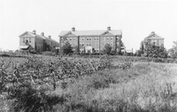 Black and white photo of a large three-story brick building with cupolas, flanked by two smaller buildings, set behind a field with rows of corn and surrounded by trees.