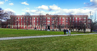 Large red-brick multi-story building with white-trimmed windows, leafless trees, and three people walking on a concrete path across a grassy lawn under a partly cloudy sky.