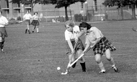 Two field hockey players fighting for possession of the ball on a grassy field. Three other players are in the background making their way toward the ball.