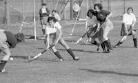 Several players are on a grassy field hockey field in the middle of a game. There are onlookers in the background.