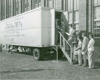 Black and white photo of a mobile X-ray unit outside a brick building, with people lined up to enter and a sign that reads 'Providence tuberculosis league's mobile x-ray unit: Who has TB? Looks won't tell but the x-ray will, play safe and have a chest x-ray.'