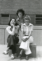 Three women posing for a photograph outside. Two are sitting on a bench while the third stands behind them.