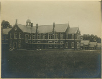 Sepia-toned photo of a large two-story brick building with a central domed tower, chimneys, and gabled roofs, set in an open grassy area with smaller buildings in the background.