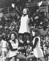 Three cheerleaders performing a stunt in front of a large crowd wearing matching uniforms with checkered patterns on the sides. Two cheerleaders are standing on the ground, supporting a third cheerleader who is elevated above them with arms raised in a 'V' shape.