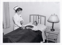 Nurse in white uniform checking the pulse of a patient lying in a hospital bed with a dark blanket, beside a table with a lamp.