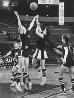 Two basketball players on a court jump for possession of the ball. Several other players are around them looking up.