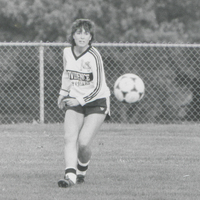 A soccer player in a Providence College uniform runs on a field with a soccer ball in the air near them.