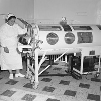 A nurse attending to a patient inside an iron lung machine, with medical equipment in the background.
