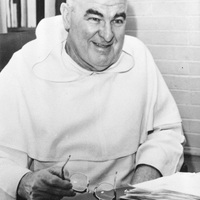 Rev. Charles V. Fennell sitting at a desk holding eyeglasses with a bookshelf in the background.
