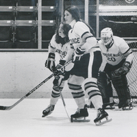 Three hockey players wearing Providence College uniforms. One stands in the goal wearing a helmet and the other two are standing on the ice with no helmets holding hockey sticks.
