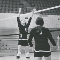 Two women wearing PC sports uniforms playing volleyball on a court in a gym. One is jumping up to hit the ball over the net.