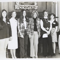 Seven women standing side by side in an indoor setting with wood-paneled walls and a framed painting above them.