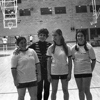 Three students in gym clothing are standing next to Helen Bert on an indoor basketball court. There are some people playing basketball in the background.