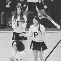 A group of cheerleaders performing at a basketball game wearing Providence College uniforms. One cheerleader is in mid-air, jumping with arms raised, while others are clapping and cheering on the court. The background shows spectators seated in bleachers