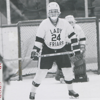 Two hockey players in Lady Friars uniforms. One is in the goal and the other skates on the ice with a hockey stick in hand.