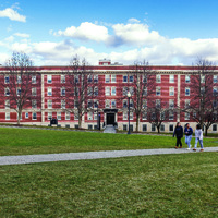 Large red-brick multi-story building with white-trimmed windows, leafless trees, and three people walking on a concrete path across a grassy lawn under a partly cloudy sky.