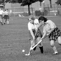 Two field hockey players fighting for possession of the ball on a grassy field. Three other players are in the background making their way toward the ball.