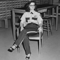A woman wearing a security guards uniform sitting on a chair with legs crossed in the Phillips Memorial Library.