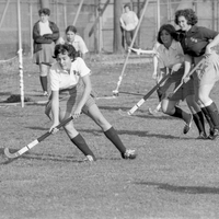 Several players are on a grassy field hockey field in the middle of a game. There are onlookers in the background.