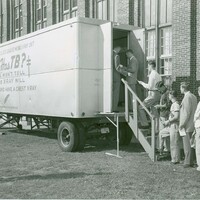 Black and white photo of a mobile X-ray unit outside a brick building, with people lined up to enter and a sign that reads 'Providence tuberculosis league's mobile x-ray unit: Who has TB? Looks won't tell but the x-ray will, play safe and have a chest x-ray.'