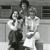 Three women posing for a photograph outside. Two are sitting on a bench while the third stands behind them.