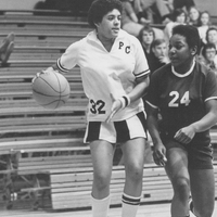 Two basketball players on the court. One has possession of the ball and is wearing a Providence College uniform. There are onlookers sitting on the bleachers in the background.