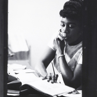 O'daylise Collins sits at a desk talking on a telephone while flipping through a notebook.