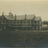 Sepia-toned photo of a large two-story brick building with a central domed tower, chimneys, and gabled roofs, set in an open grassy area with smaller buildings in the background.