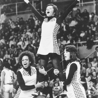 Three cheerleaders performing a stunt in front of a large crowd wearing matching uniforms with checkered patterns on the sides. Two cheerleaders are standing on the ground, supporting a third cheerleader who is elevated above them with arms raised in a 'V' shape.