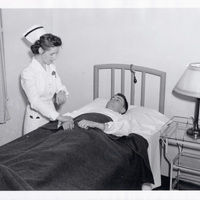 Nurse in white uniform checking the pulse of a patient lying in a hospital bed with a dark blanket, beside a table with a lamp.