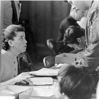 Sally J Thibodeau sitting at a registration desk with papers in front of her, interacting with a man standing on the other side of the desk.