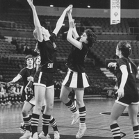 Two basketball players on a court jump for possession of the ball. Several other players are around them looking up.